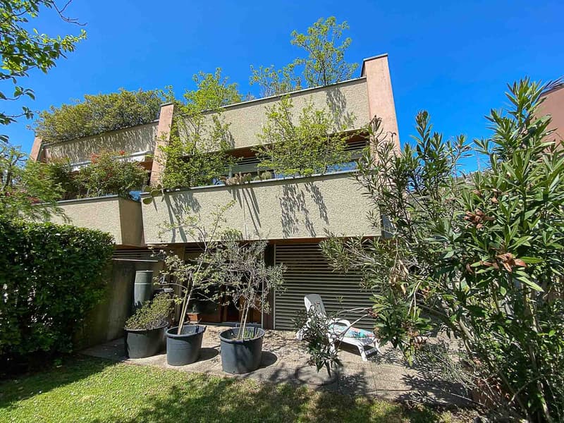 A two-story house with trees and plants, a balcony, a chair, and some pots on the ground