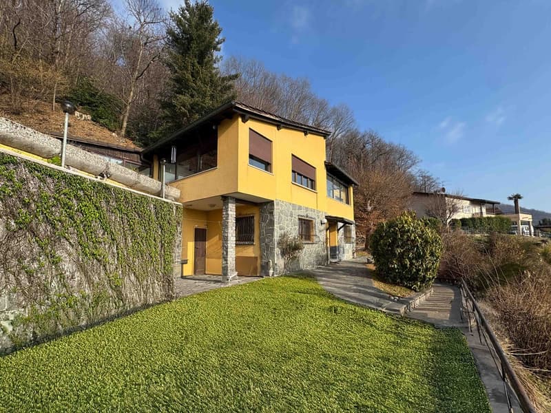 Yellow house with stone walls, raised ground floor, large windows, green lawn, stone pathway, trees, mountain view