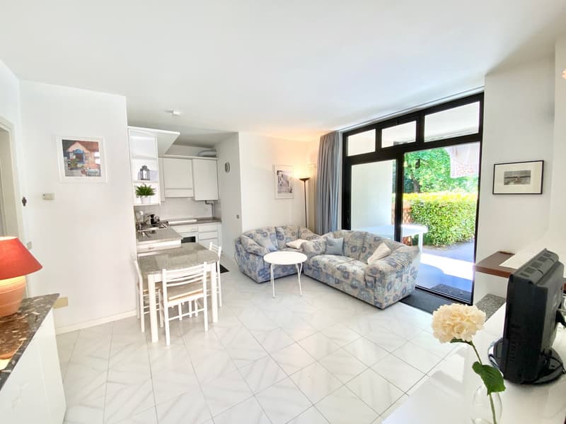 Open-plan living room and kitchen, large windows leading to a balcony, tiled flooring, floral patterned sofa, coffee table, and wall decor.