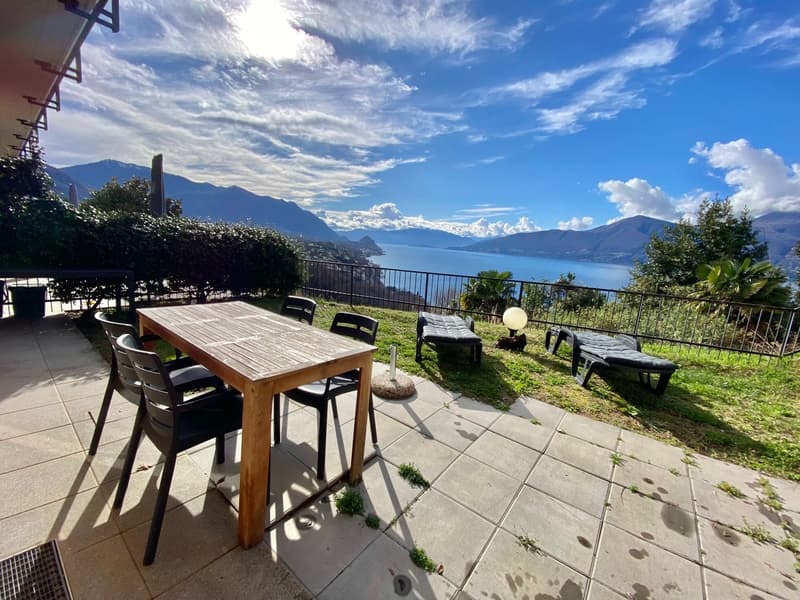 Balcony with wooden table, 4 chairs, 2 sunbeds, lamp, green plants, fence, scenic lake view