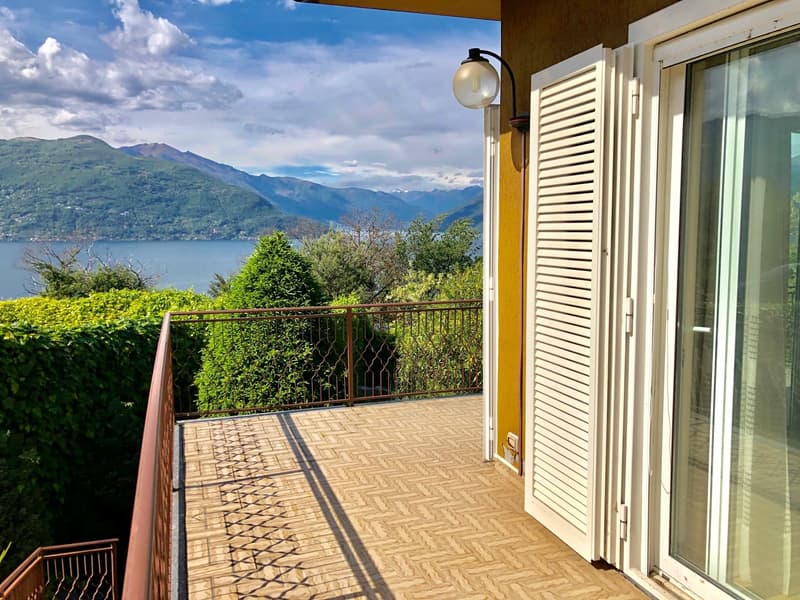 Spacious balcony with a stunning view of the lake and mountains. The balcony has a paved floor and a metal railing. There is a lamp fixture on the wall and the balcony is surrounded by lush greenery.