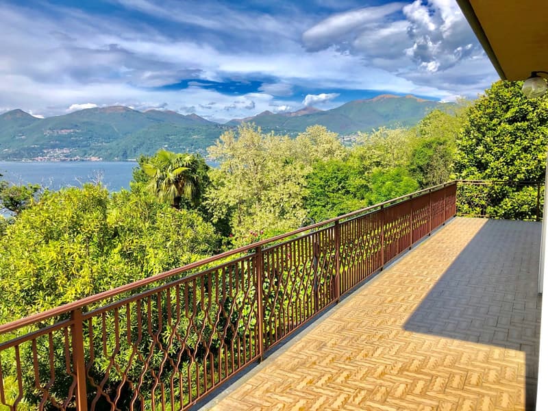 A wooden terrace with a metal railing overlooking a lush, green landscape. The terrace offers a panoramic view of mountains, a lake, and a town in the distance. The surrounding vegetation includes palm trees and other foliage, creating a serene and pictur