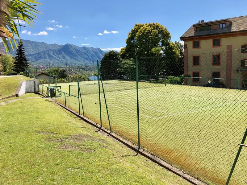 The image shows a tennis court surrounded by a green wire fence. The court has a green surface and is located in a scenic outdoor setting with mountains and trees in the background.