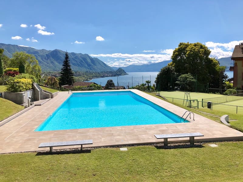 The image shows a large outdoor swimming pool surrounded by a well-manicured lawn and lush greenery. The pool is situated in a scenic location, with mountains and a lake visible in the background. There are also some benches and a tennis court visible in