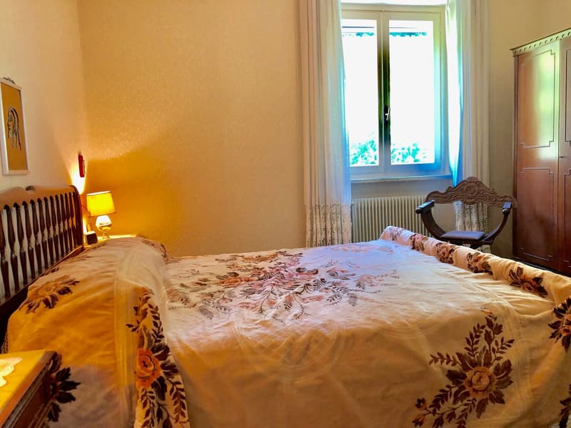 Spacious bedroom with a large bed, floral patterned bedding, and a wooden armoire. The room has warm, yellow-toned walls and a large window that provides natural light.
