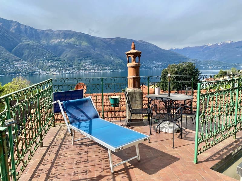 Outdoor terrace with a scenic view of a lake surrounded by mountains. The terrace has a lounge chair, a table with chairs, and a decorative chimney-like structure. The terrace is enclosed by a wrought-iron railing and offers a panoramic view of the lake a