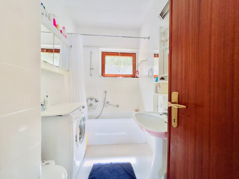 Spacious bathroom with a washing machine, sink, and shower. The room has white walls and tile flooring, with a window providing natural light.