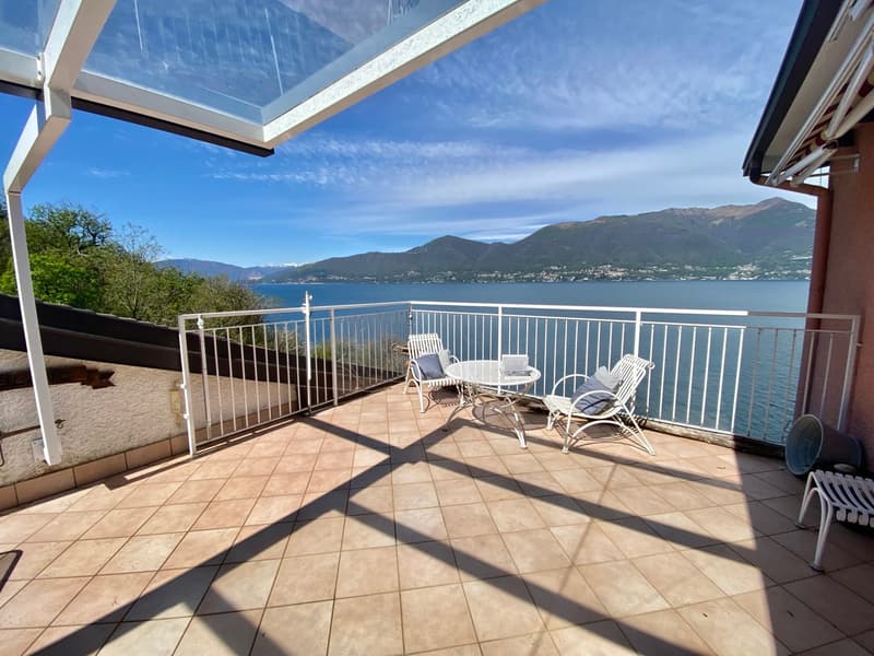 Spacious balcony with tiled flooring, white railings, and outdoor furniture including chairs and a table. The balcony offers a stunning view of the lake and mountains in the background.
