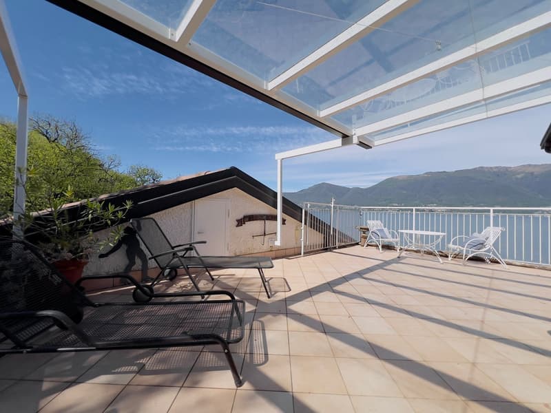 Spacious outdoor terrace with glass roof, providing a panoramic view of the surrounding mountains. The terrace features comfortable patio furniture, including lounge chairs and a table, as well as potted plants, creating a relaxing and scenic environment.