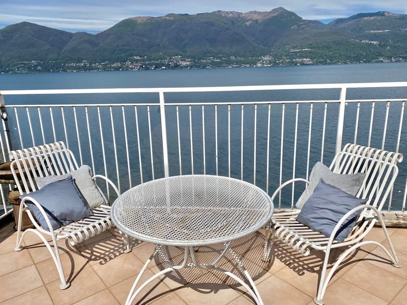 Outdoor balcony with a round metal table and two rocking chairs, overlooking a scenic lake surrounded by mountains