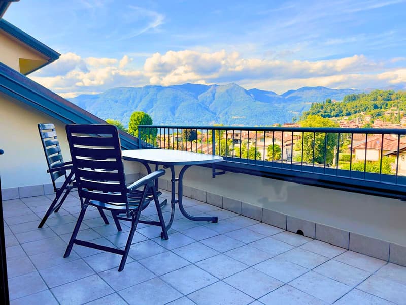 Outdoor balcony with a table and chairs, overlooking a scenic mountain landscape with blue skies and clouds