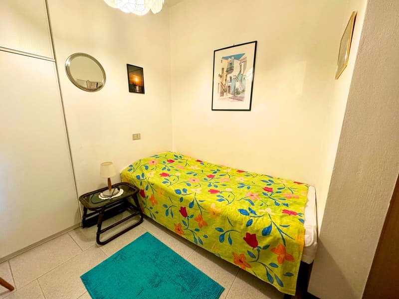 Bedroom with a floral-patterned bedspread, a side table with a lamp, a framed artwork on the wall, and a round mirror.