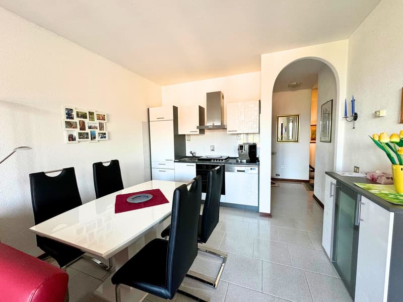The image shows an open-concept kitchen and dining area. The kitchen features white cabinets, a stainless steel oven and range hood, and a tiled backsplash. The dining area has a white table with black chairs. The room has tile flooring throughout and whi