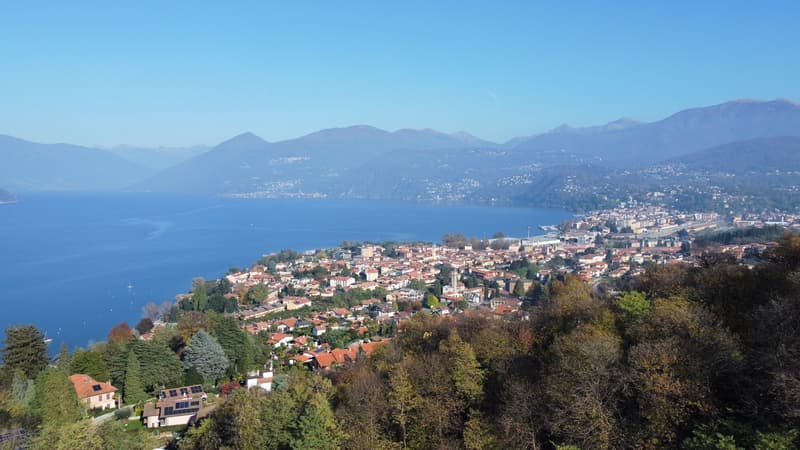 Aerial view of a lakeside town surrounded by mountains. The town has a mix of residential and commercial buildings with red-tiled roofs. The lake is a deep blue color and the mountains in the background are hazy. There are trees and greenery throughout th