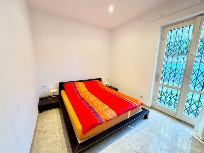 Spacious bedroom with white walls, large window with decorative grille, tiled floor, and a modern black bed frame with a vibrant red and orange striped comforter.