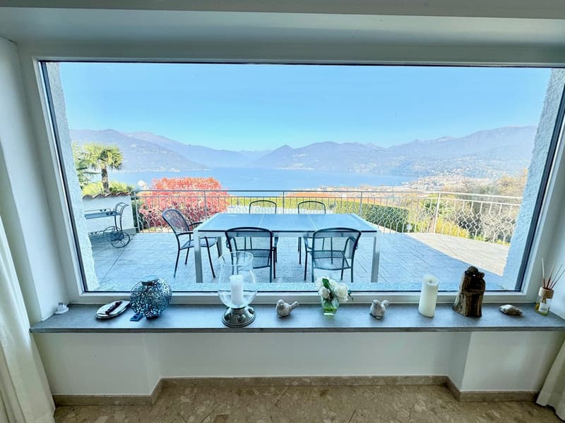 Large windows with a scenic view of the mountains and lake, outdoor dining area with a table and chairs on a balcony or terrace, decorative items on the windowsill including candles, rocks, and a glass vase