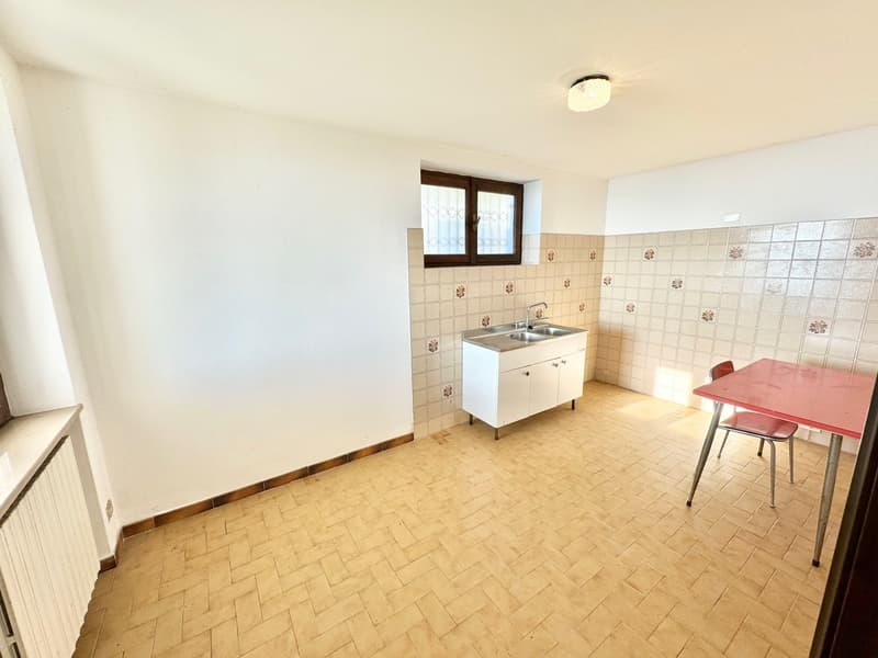 An empty kitchen with tiled floor, white walls, double sink, red table, red chair, small window, and radiator on the wall.