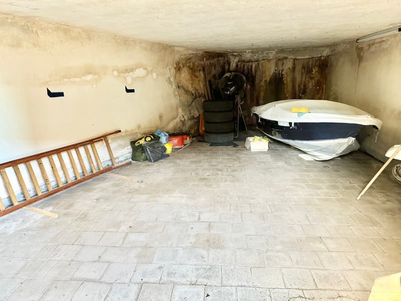 The image shows a basement or underground storage space. The room has a concrete floor with tiled flooring, and the walls appear to be made of stone or concrete. There are various items stored in the room, including a ladder, some bags, and what looks lik