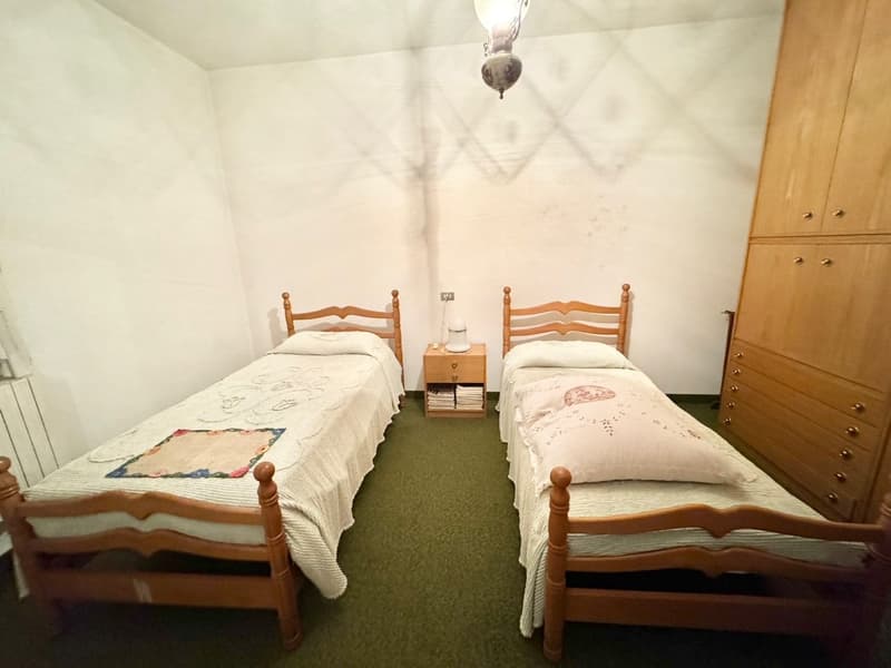 The image shows a bedroom with two twin beds, each with a wooden headboard. There is a wooden dresser between the beds, and a hanging light fixture above. The room has white walls and a green carpeted floor.