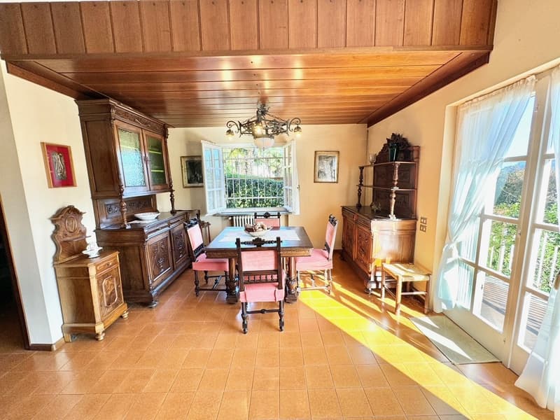Spacious dining room with wooden paneled ceiling, large windows, antique wooden furniture including a dining table with chairs, a sideboard, and a display cabinet. The room has a cozy and rustic feel.