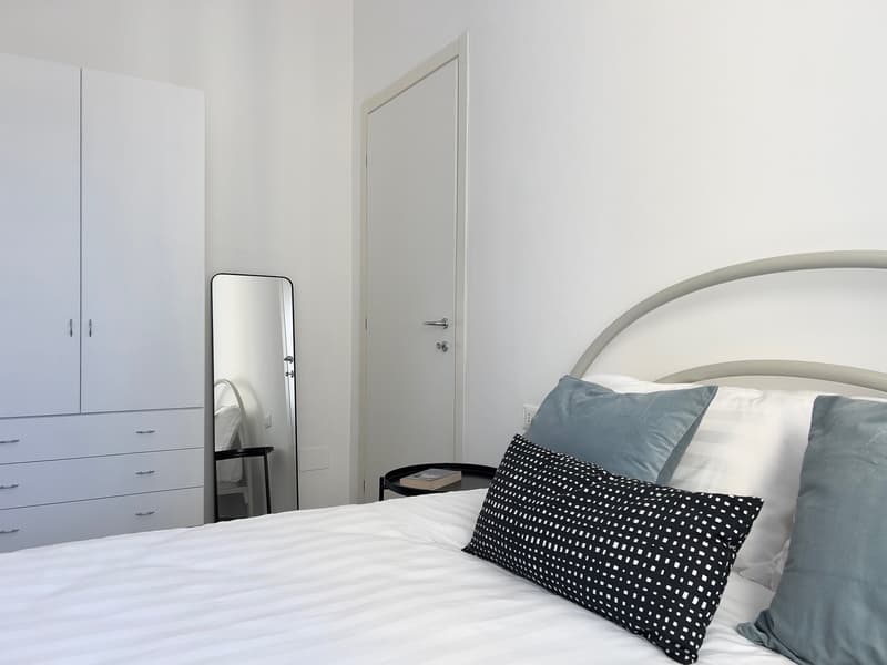 Large bed with white sheets and pillows, a white wardrobe, a mirror and a nightstand with a book on it