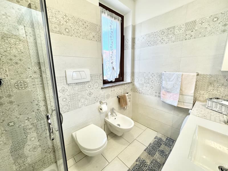 White tiles, glass shower door, white sink, toilet, bidet, towels, rug, toilet paper