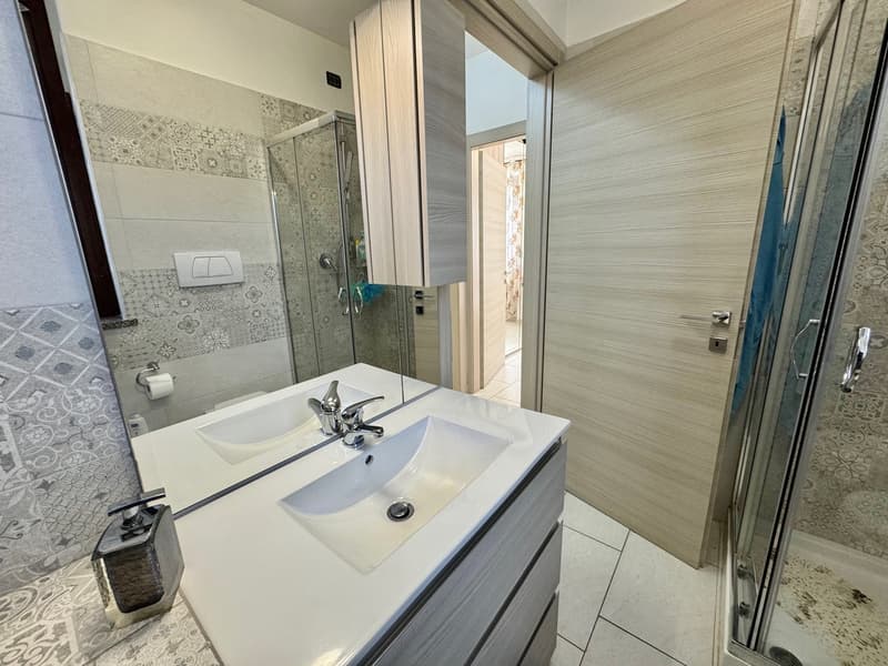 modern bathroom, white walls, white tiles, silver fixtures, glass shower door, toilet, white sink, vanity cabinet, mirror, floor soap dispenser