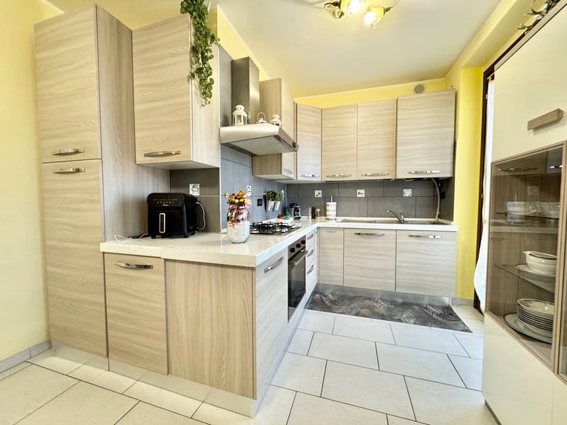 The image shows a modern kitchen with light-colored wood cabinets, a white countertop, and a tiled floor. The kitchen is equipped with a dishwasher, a coffee maker, and other small appliances. The walls are painted in a light yellow color, and there is a