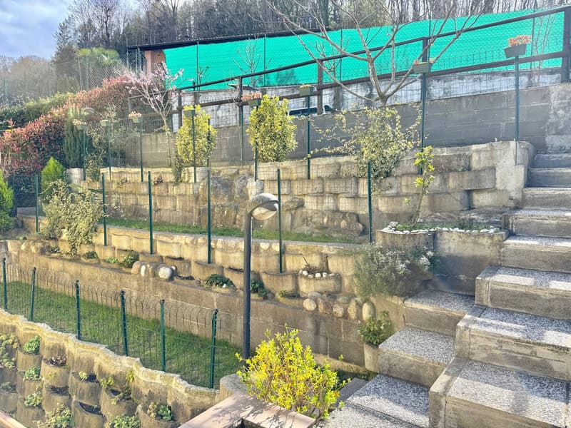 Terraced garden with multiple layers, each featuring plants and trees, stairs for access, stone walling, net fencing, and lamp posts.