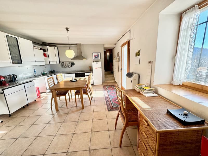 The image shows an open-plan kitchen and dining area. The kitchen features white cabinets, a stainless steel range hood, and a dishwasher. There is a wooden dining table with chairs in the center of the room. The room has tile flooring and large windows p