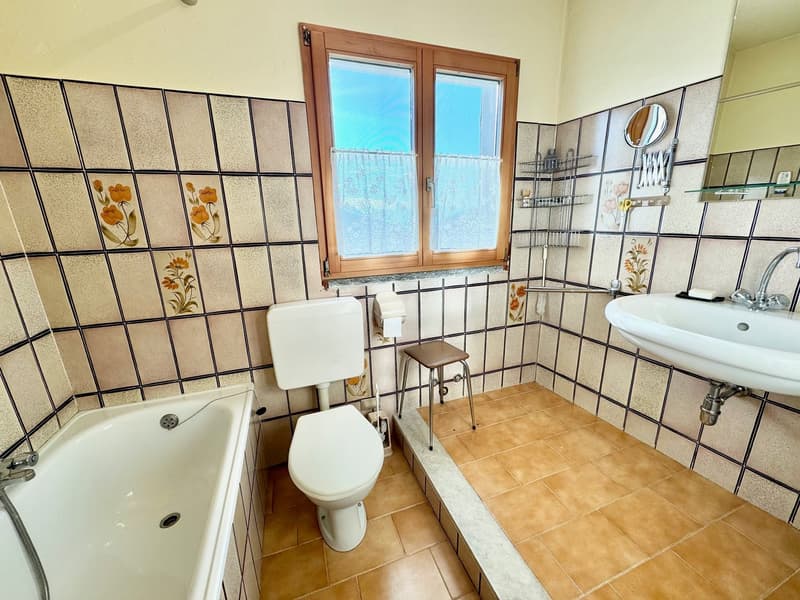 Bathroom with bathtub, toilet, sink, window, tiles, toiletries, stool, and a mirror.