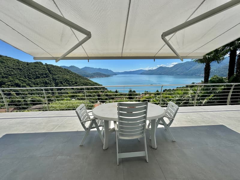 Covered outdoor dining area with a round table and chairs, overlooking a scenic lake surrounded by mountains and lush greenery.