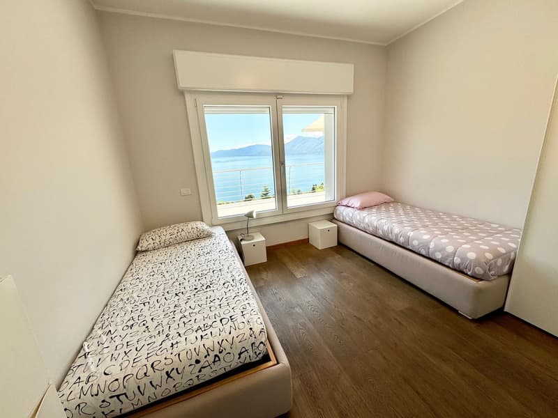 Spacious bedroom with large windows offering a scenic view, hardwood flooring, two single beds with patterned bedding, and a side table with a lamp.