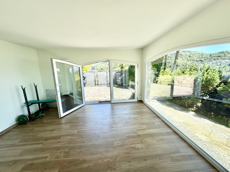 Spacious living room with large windows and sliding glass doors leading to a balcony or terrace. The room has hardwood flooring and a view of the surrounding greenery outside.