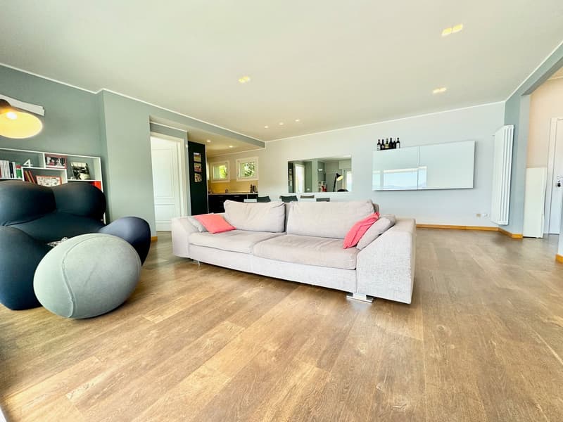 Spacious living room with hardwood flooring, light gray walls, large sofa with colorful pillows, and a bookshelf. The room has large windows that provide natural light.