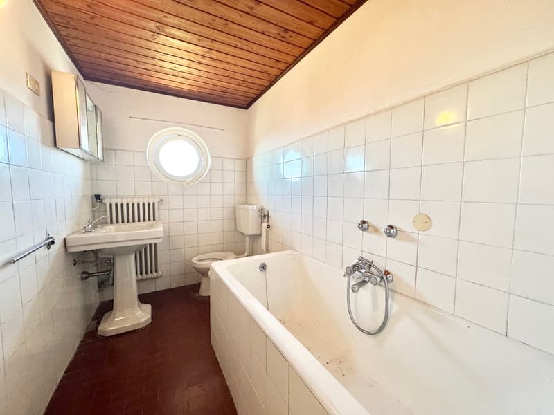 The image shows a bathroom with a white tiled walls, a pedestal sink, a toilet, and a clawfoot bathtub with a shower fixture. The ceiling has a wooden paneled design. The bathroom appears to be in an older style or vintage setting.