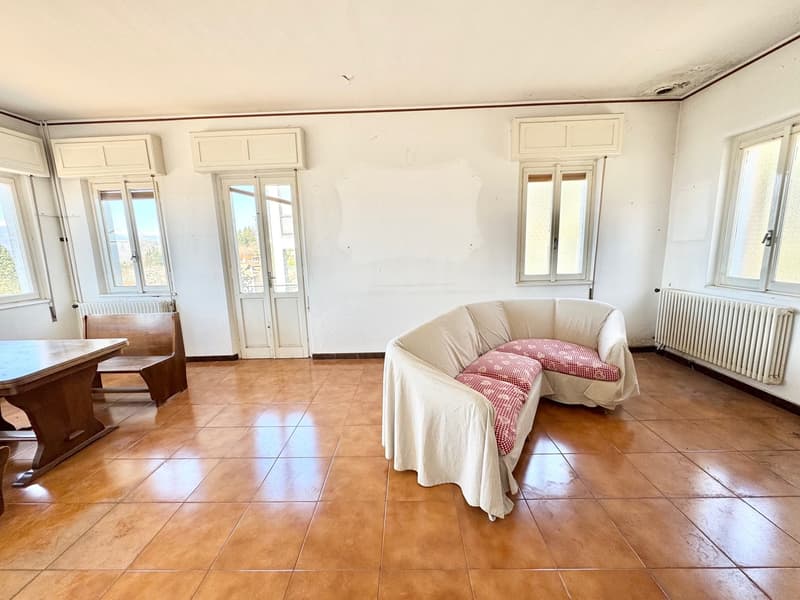 Spacious living room with large windows, hardwood floors, and a couch with a checkered blanket. The room also has a wooden table and chairs.