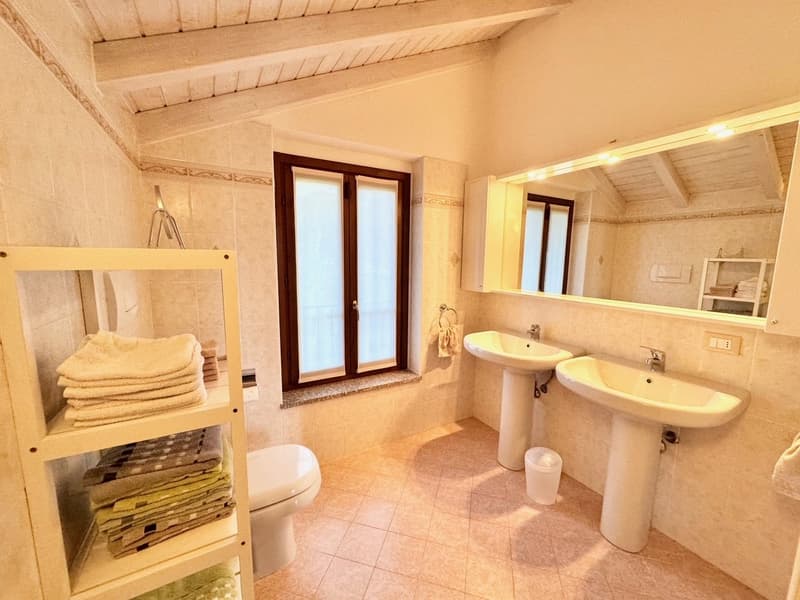 Spacious bathroom with tiled floor, double sinks, toilet, and a towel rack. The room has large windows and a wooden ceiling with exposed beams.