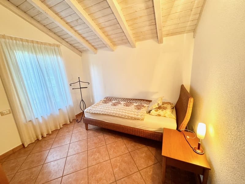 Bedroom with wooden ceiling beams, tiled floor, curtains, bed with patterned bedding, and a decorative cross on the wall.