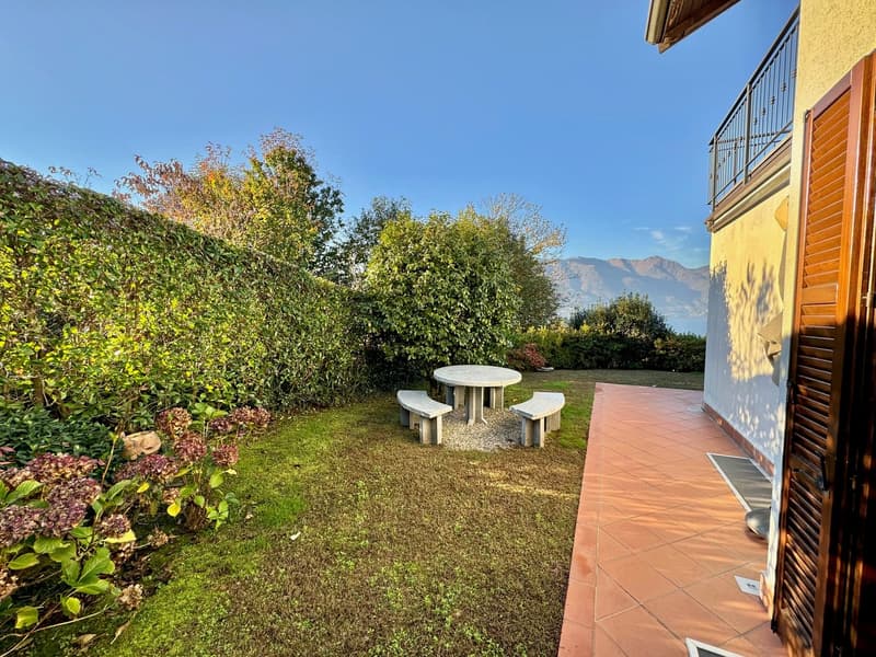 The image shows a well-maintained outdoor area with a grassy lawn, a stone table and benches, and lush vegetation surrounding the space. The area appears to be part of a larger property, with a building visible in the background and a balcony or terrace o