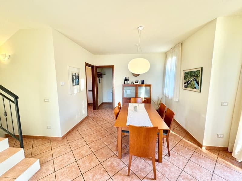 The image shows a spacious dining room with a wooden dining table and chairs. The room has tile flooring and white walls, with a wooden cabinet and artwork on the walls. There is a doorway visible leading to another room.