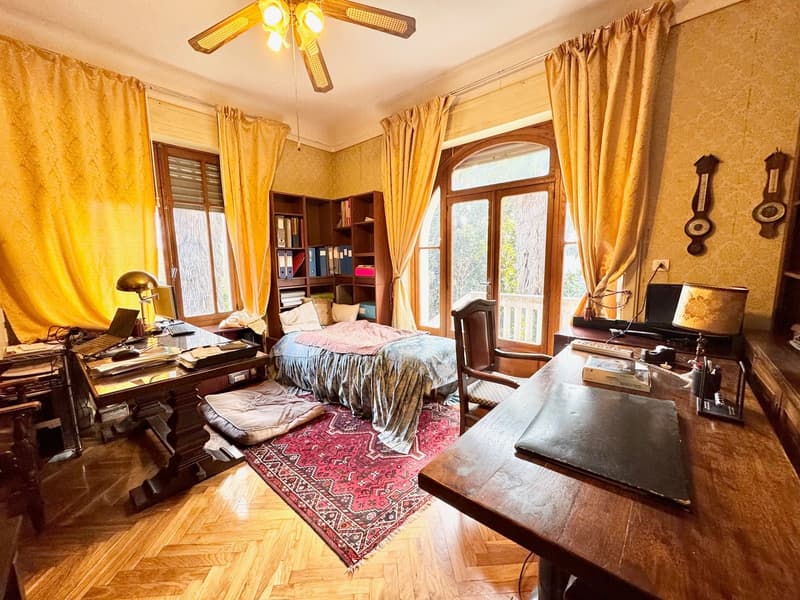 Spacious bedroom with hardwood floors, large windows, yellow curtains, a bed with patterned bedding, a wooden desk, bookshelves, and a record player.