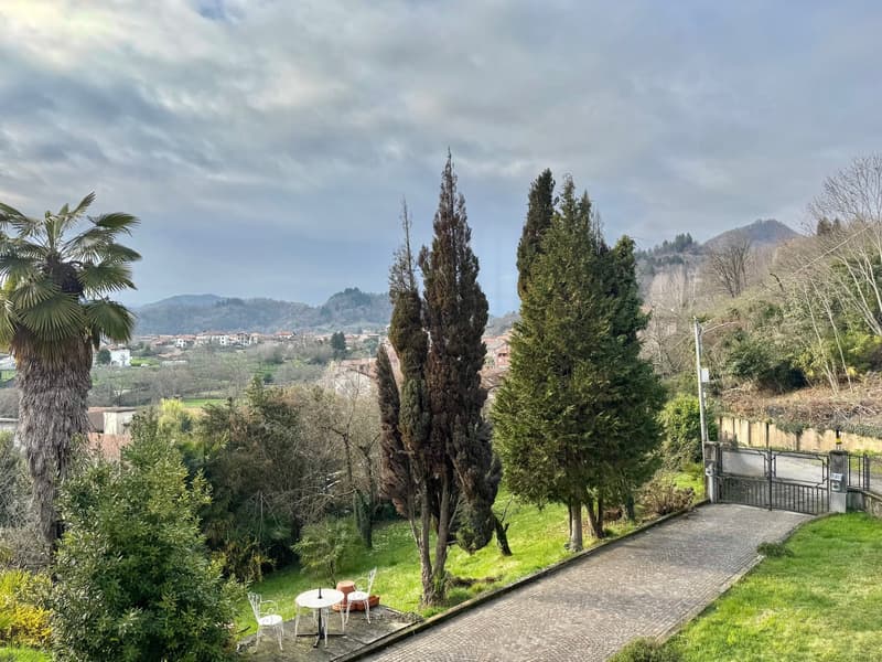The image shows a scenic landscape with a view of a small town or village nestled among rolling hills and mountains. The foreground features a paved walkway surrounded by lush greenery, including palm trees, pine trees, and other vegetation. In the backgr