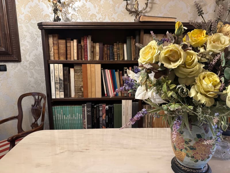 Wooden bookshelf with various books, a marble-topped table, and a vase of yellow roses and other flowers on the table.