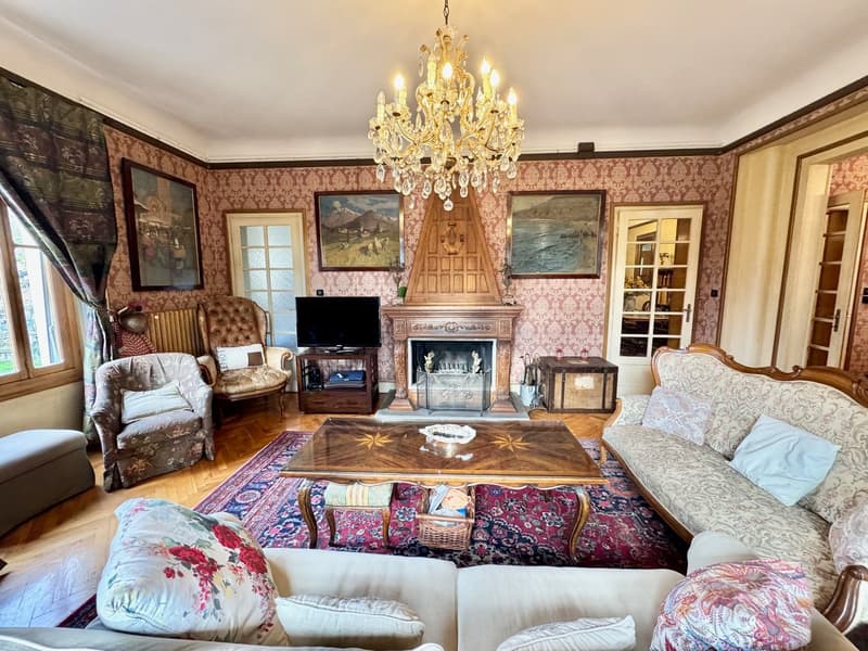 Ornate living room with floral wallpaper, crystal chandelier, tufted leather armchair, wooden coffee table, and a fireplace with a decorative mantel