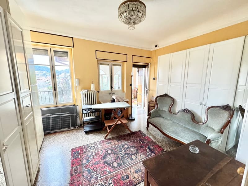 Spacious living room with large windows, ornate chandelier, antique furniture including a sofa and armchairs, a wooden table, and a Persian-style rug on the floor.