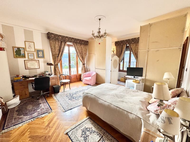 Spacious bedroom with hardwood floors, large windows, a chandelier, a desk, a pink armchair, and a bed with a patterned rug in front of it.