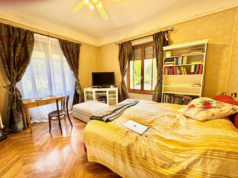 Spacious bedroom with yellow walls, large windows, wooden floor, a bed with yellow and patterned bedding, a TV stand, a bookshelf, and a desk with a chair.