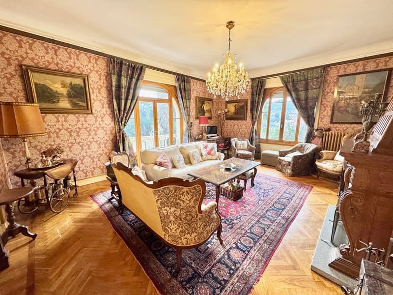 Spacious living room with ornate wallpaper, large windows, a grand chandelier, antique furniture including a sofa, armchairs, and a coffee table, and a decorative fireplace