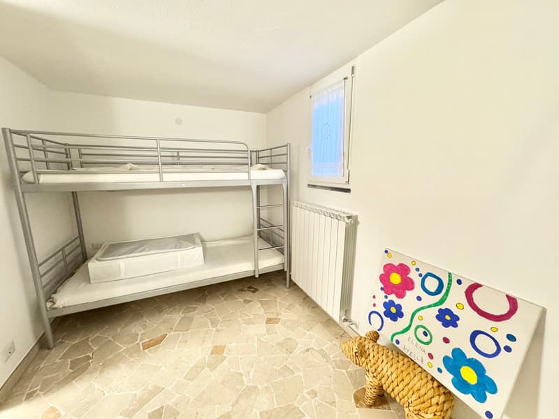 The image shows a bedroom with a bunk bed, a radiator, and a colorful painting on the wall. The floor is made of stone tiles.